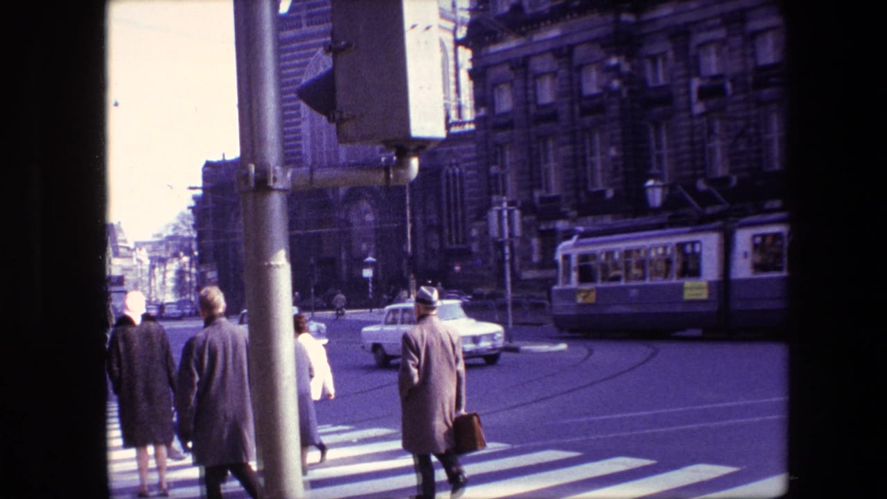 Amsterdam, 1967 intersection view with pedestrians crossing in coats, a traffic signal pole in the foreground, a tram at right, and a white car on the street.