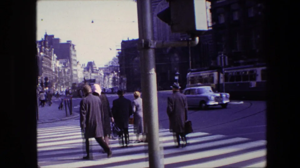Amsterdam, 1967 view down a city street as pedestrians cross a zebra crossing while a white car and tram pass near historic multi-story buildings.