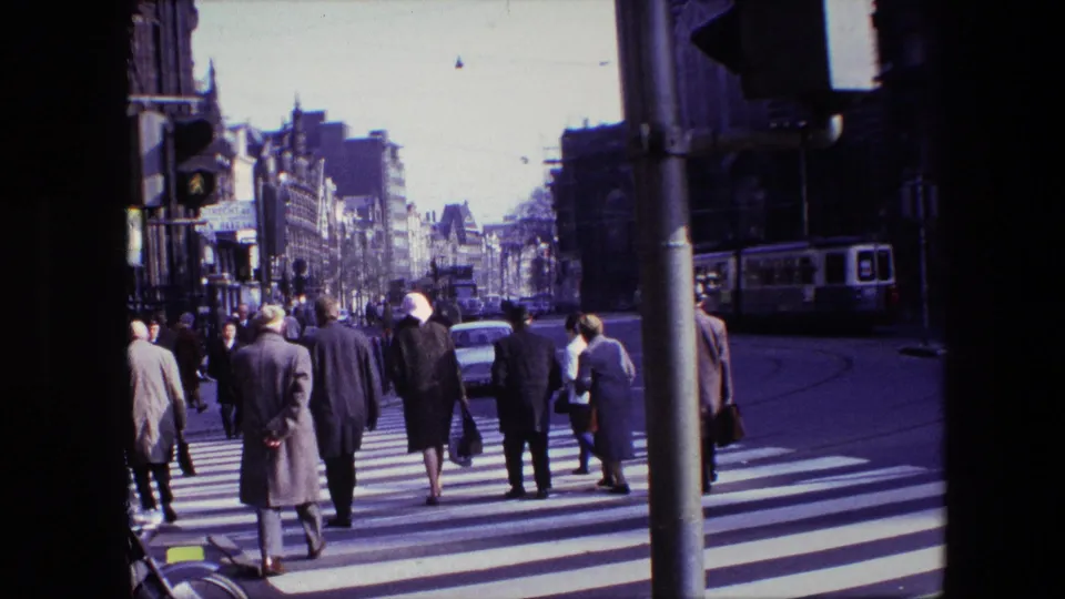 Amsterdam, 1967 urban crossing with a group of pedestrians seen from behind, tram tracks on the roadway, cars ahead, and a tram along the right side of the frame.
