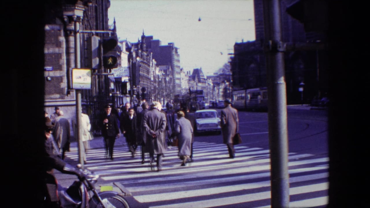 Cataloged Amsterdam 1967 frame with pedestrians crossing a zebra crossing, a white car on the road, a traffic signal, bicycles, and dense urban facades in view.