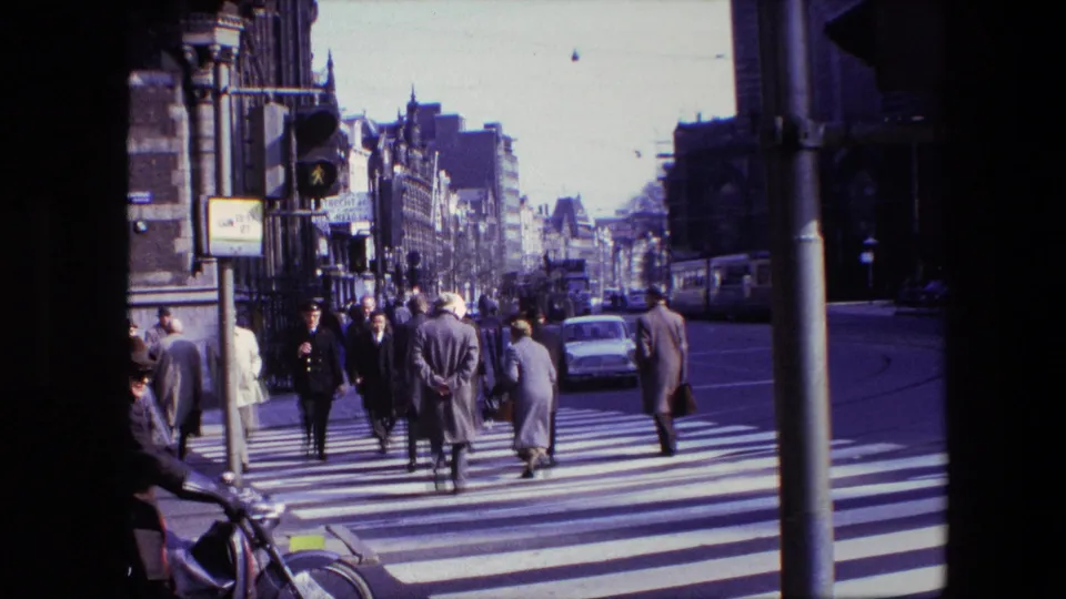 Cataloged Amsterdam 1967 frame with pedestrians crossing a zebra crossing, a white car on the road, a traffic signal, bicycles, and dense urban facades in view.