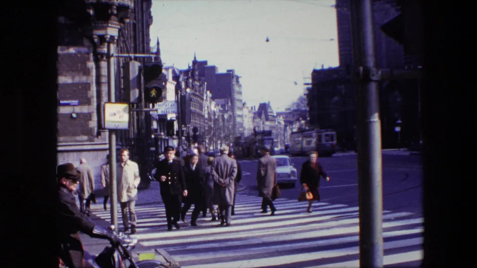 Cataloged Amsterdam 1967 street scene showing a uniformed officer among pedestrians on a zebra crossing, with a traffic light, bicycles, a foreground rider, and urban buildings behind.