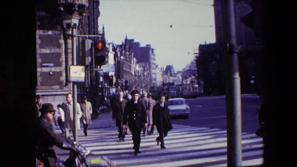 Cataloged Amsterdam 1967 frame with pedestrians finishing a zebra crossing under a red traffic signal, with a white car, bicycles, street poles, and tall urban buildings visible.