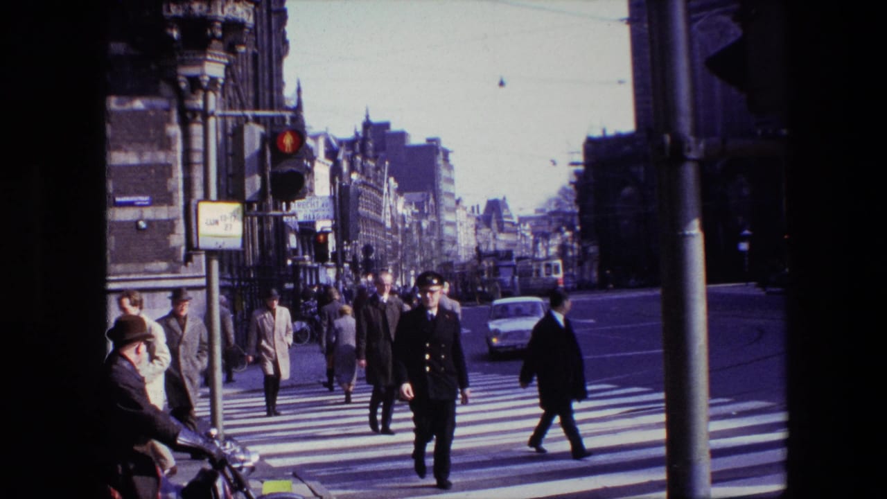 Amsterdam street scene from the 1967 clip showing pedestrians, a uniformed traffic officer, classic cars, sidewalks, and overhead tram wires near a zebra-striped crosswalk.