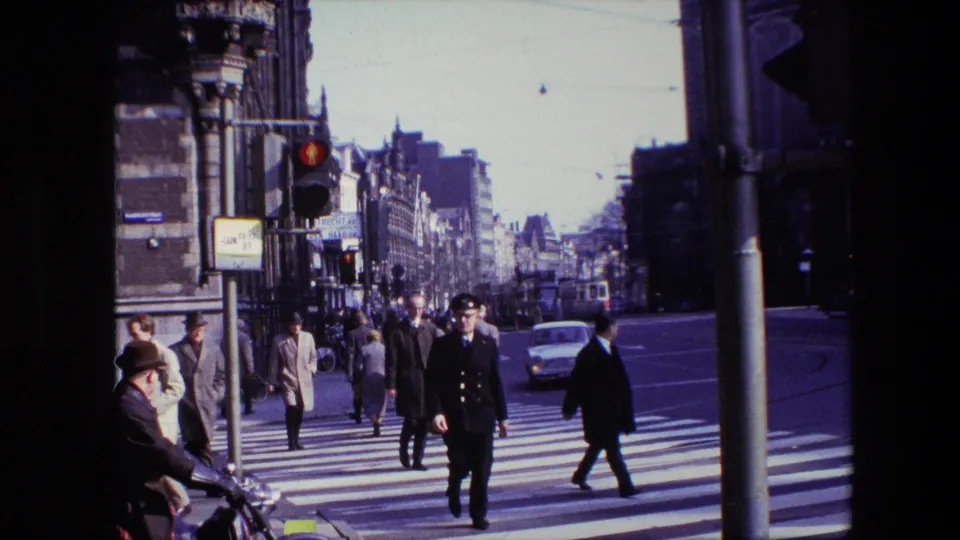 Amsterdam street scene from the 1967 clip showing pedestrians, a uniformed traffic officer, classic cars, sidewalks, and overhead tram wires near a zebra-striped crosswalk.