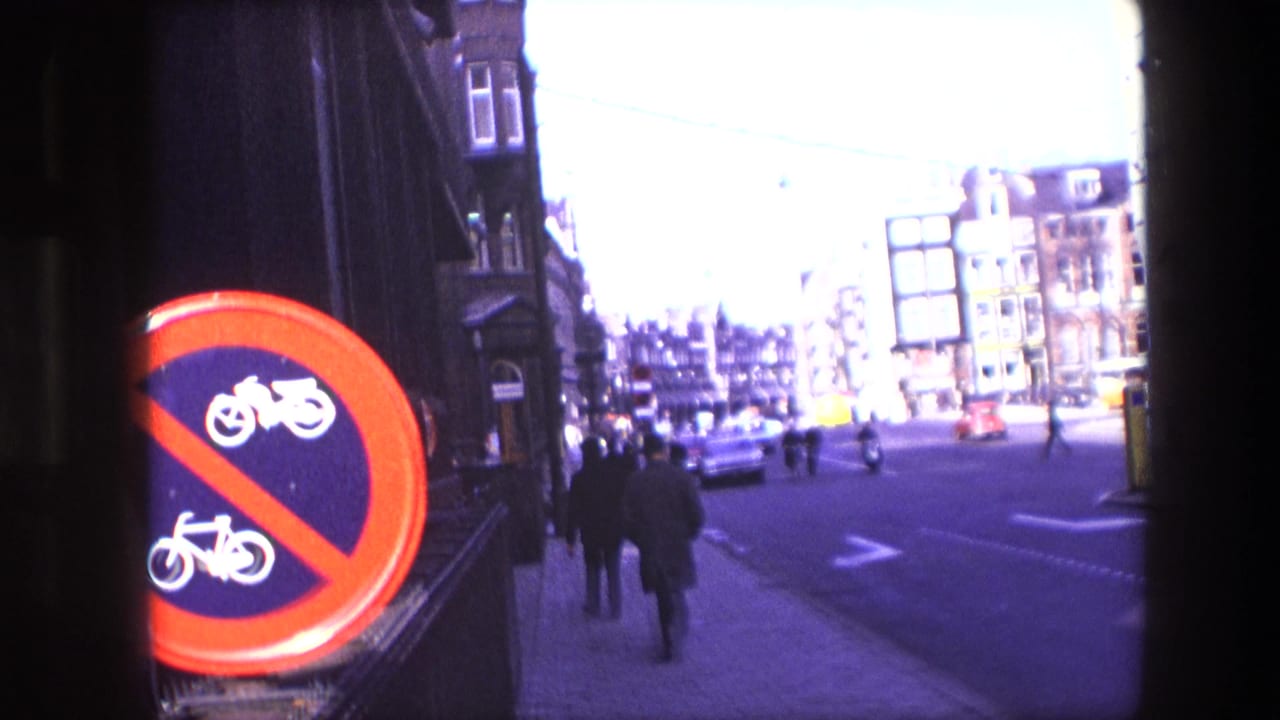 Amsterdam street view from the 1967 clip with a prominent red bicycle and motorcycle restriction sign, pedestrians, cars, scooters, overhead wires, and tall urban buildings.