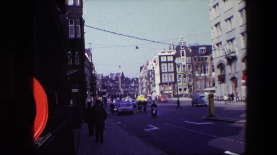 Amsterdam street scene from the 1967 clip showing scooters and cars moving through an intersection while pedestrians walk along the sidewalk beneath overhead tram wires.