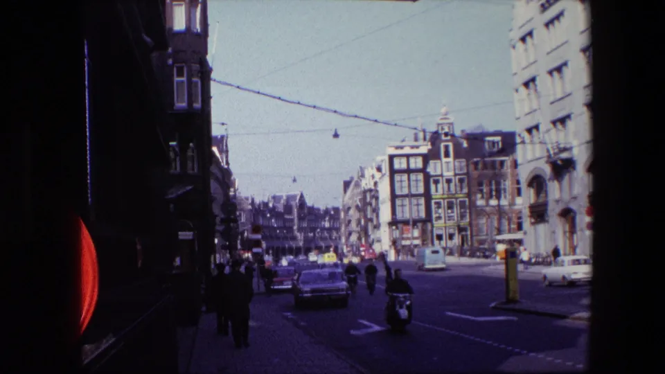 Amsterdam street view from the 1967 clip showing a scooter rider, cars, pedestrians on the sidewalk, overhead tram wires, and narrow multi-story buildings at a busy intersection.