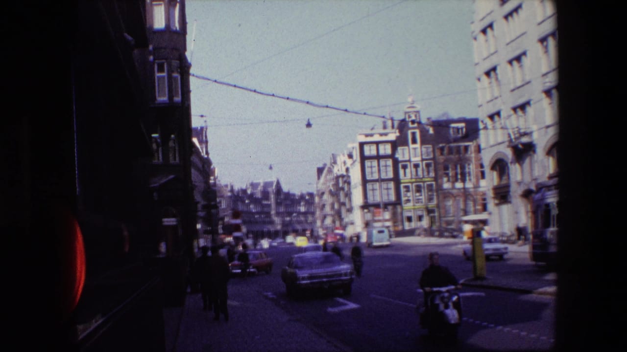 1967 Amsterdam street view with classic cars, pedestrians, cyclists, overhead tram wires, and narrow buildings seen from a shaded curbside position.