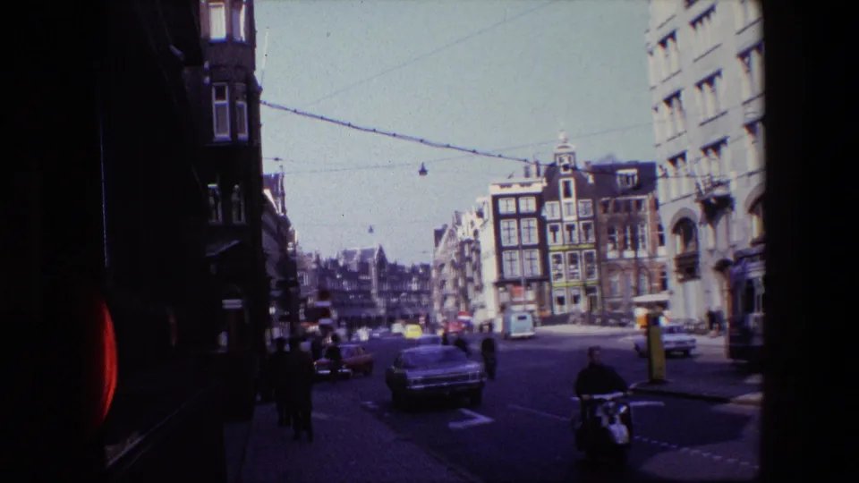 1967 Amsterdam street view with classic cars, pedestrians, cyclists, overhead tram wires, and narrow buildings seen from a shaded curbside position.