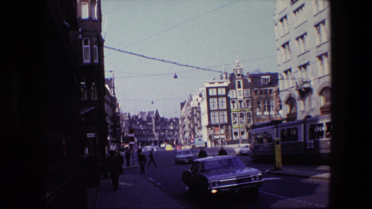 Amsterdam intersection in 1967 with pedestrians crossing near classic cars, overhead tram wires, and multi-story streetfront buildings.
