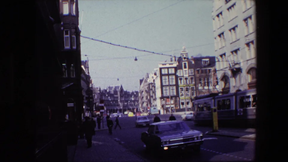 Amsterdam intersection in 1967 with pedestrians crossing near classic cars, overhead tram wires, and multi-story streetfront buildings.