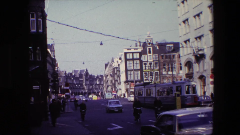 1967 Amsterdam street scene with a tram passing at the right edge, cars and cyclists in traffic, pedestrians on the sidewalk, and overhead transit wires.