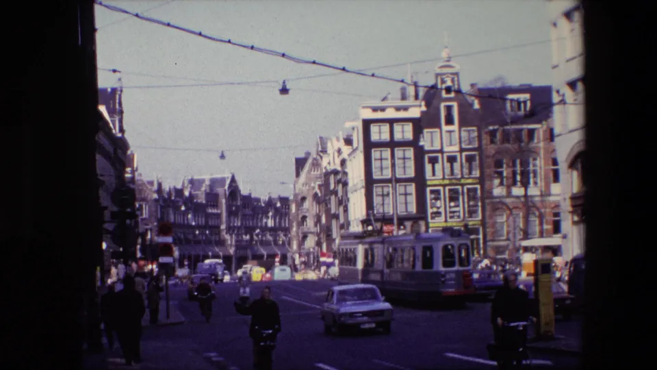 1967 Amsterdam street view with a tram alongside cars and cyclists, pedestrians near the curb, overhead wires, and older narrow urban buildings in sunlight.