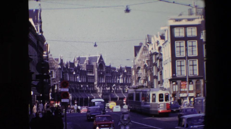 Amsterdam intersection in 1967 with a red-and-white tram, compact cars, cyclists, pedestrians, suspended street lamps, and historic facades.