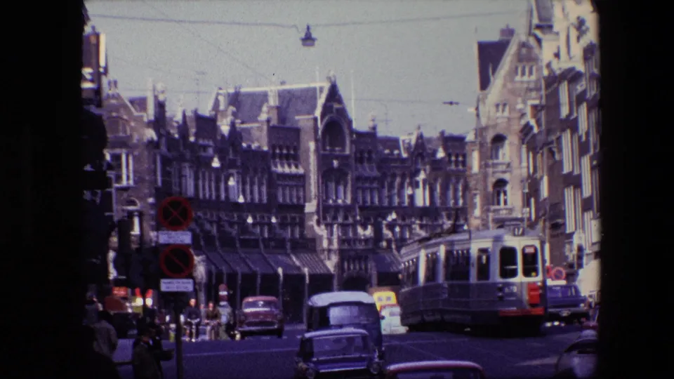 1967 Amsterdam street scene with period cars in the foreground, an electric tram to the right, pedestrians, traffic signs, and ornate building facades.