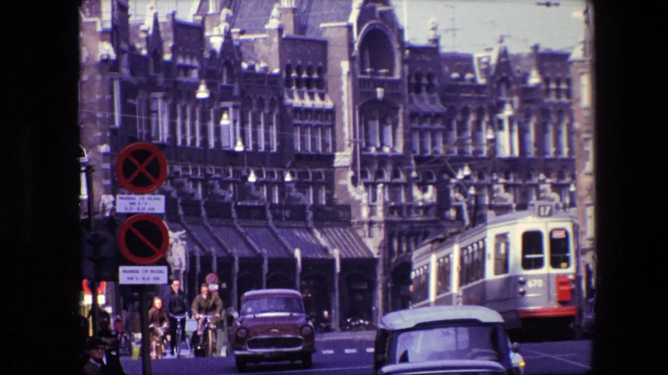 Amsterdam street traffic in 1967 with cyclists near the center, a compact car and covered vehicle in the foreground, a tram at right, and historic architecture behind them.