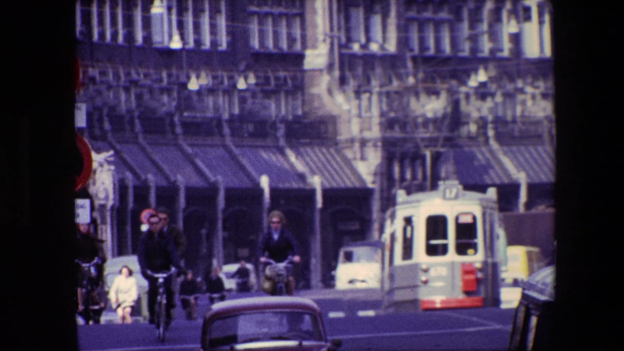 Bicyclists ride along a broad street as a tram and cars pass near storefronts.