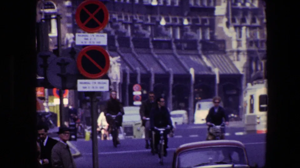 Cyclists ride toward the camera on a busy Amsterdam street in 1967, with classic vehicles, no-parking signs, traffic signals, and storefronts visible.