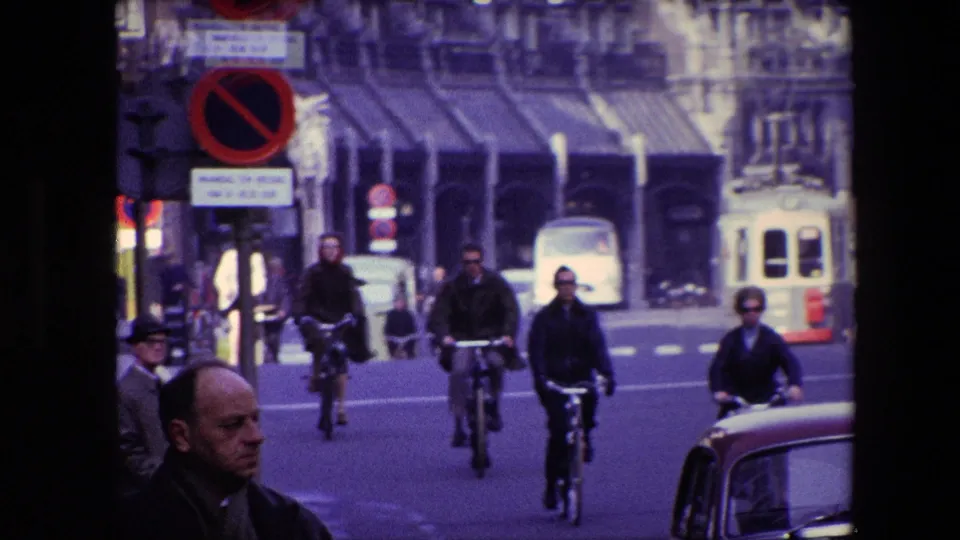 End frame of the 1967 Amsterdam street clip, with multiple cyclists in dark clothing, a foreground car, a tram in the distance, and pedestrians along the road.