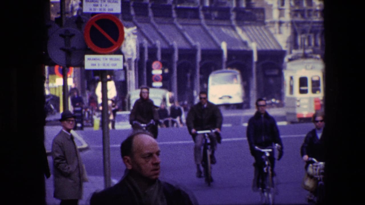 1967 Amsterdam street scene with adult cyclists approaching the camera, pedestrians near the curb, classic vehicles, road signs, and ornate building facades in the background.