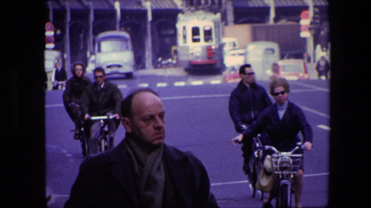 1967 Amsterdam street view with a man in the foreground, cyclists riding behind him, classic cars, a light-colored tram or bus, and multi-story urban buildings.