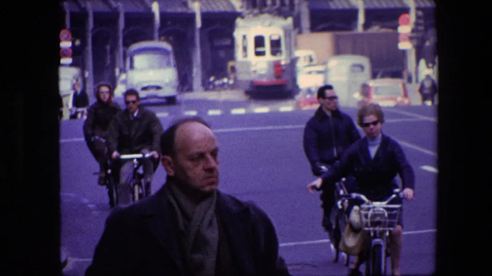 1967 Amsterdam street view with a man in the foreground, cyclists riding behind him, classic cars, a light-colored tram or bus, and multi-story urban buildings.