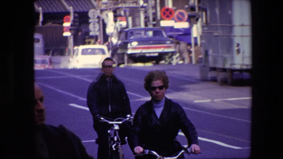 1967 Amsterdam footage showing two adult cyclists in sunglasses riding toward the camera, with classic cars, traffic signs, roadway lanes, and urban buildings behind them.