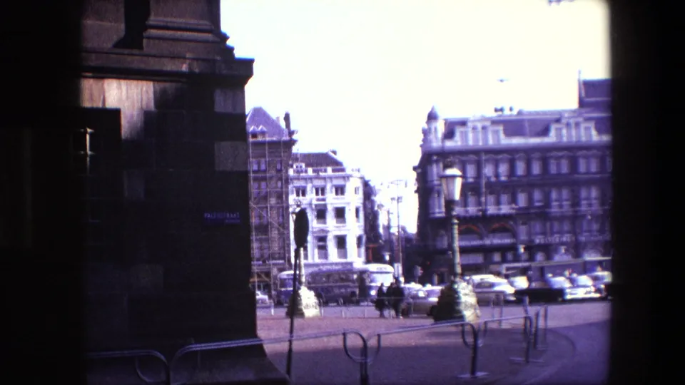 1967 Amsterdam street corner framed beside a dark building edge, showing distant pedestrians, cars, multi-story facades, street furniture, and a bright overexposed sky.