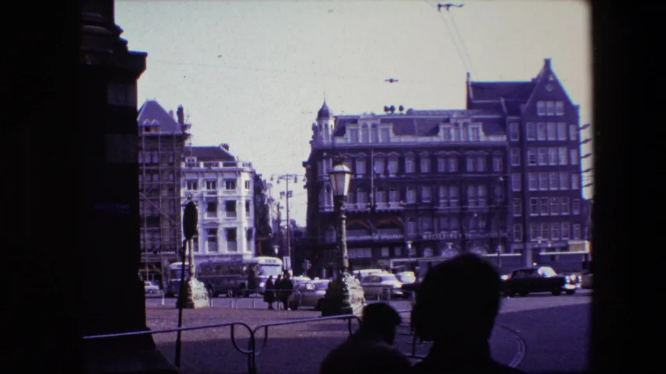 A close foreground silhouette passes through the shaded edge of a 1967 Amsterdam square view, while cars, pedestrians, tram wires, lamps, and older buildings remain visible in the distance.
