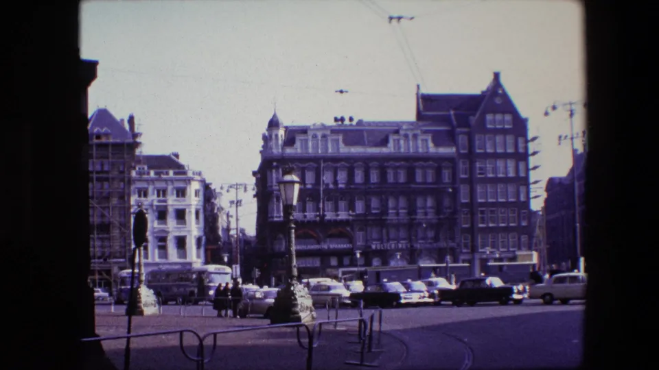 The final frame holds on a 1967 Amsterdam street square with moving cars, distant pedestrians, overhead tram wires, lamps, and historic facades seen through a dark structural opening.