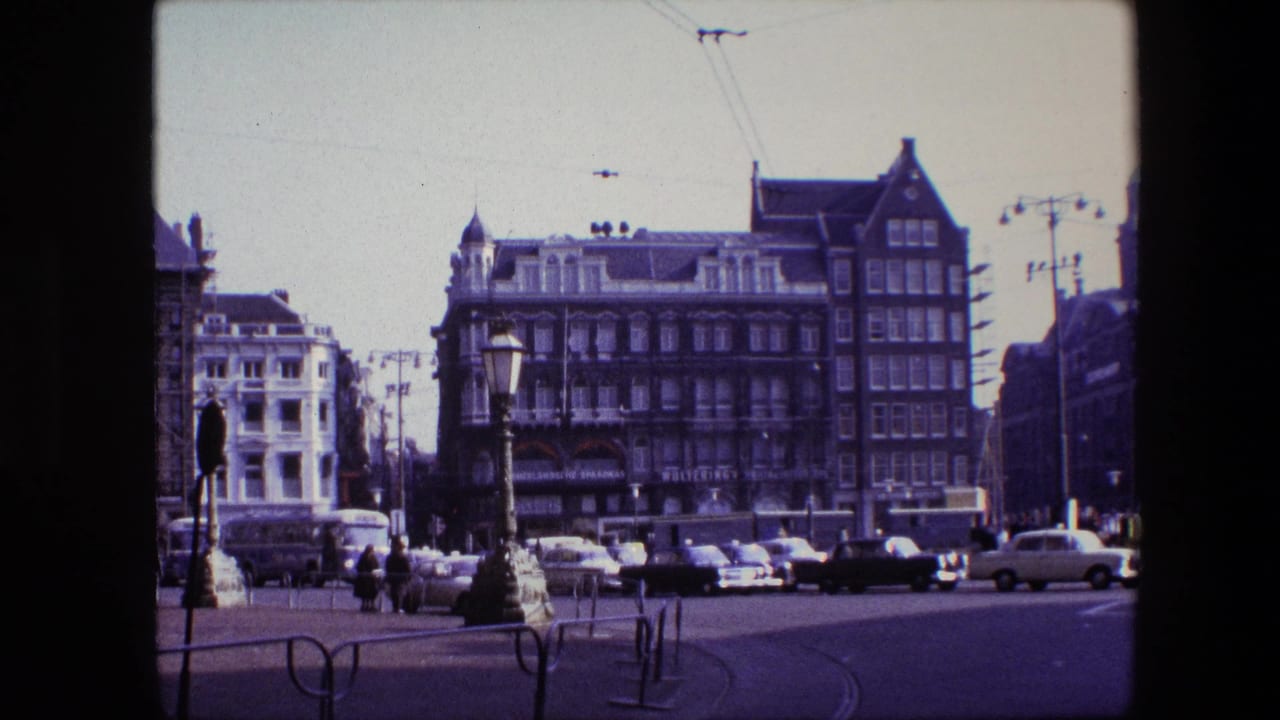 1967 Amsterdam street intersection with cars, pedestrians, overhead tram wires, and gabled commercial buildings seen from a stationary 8mm home-movie viewpoint.