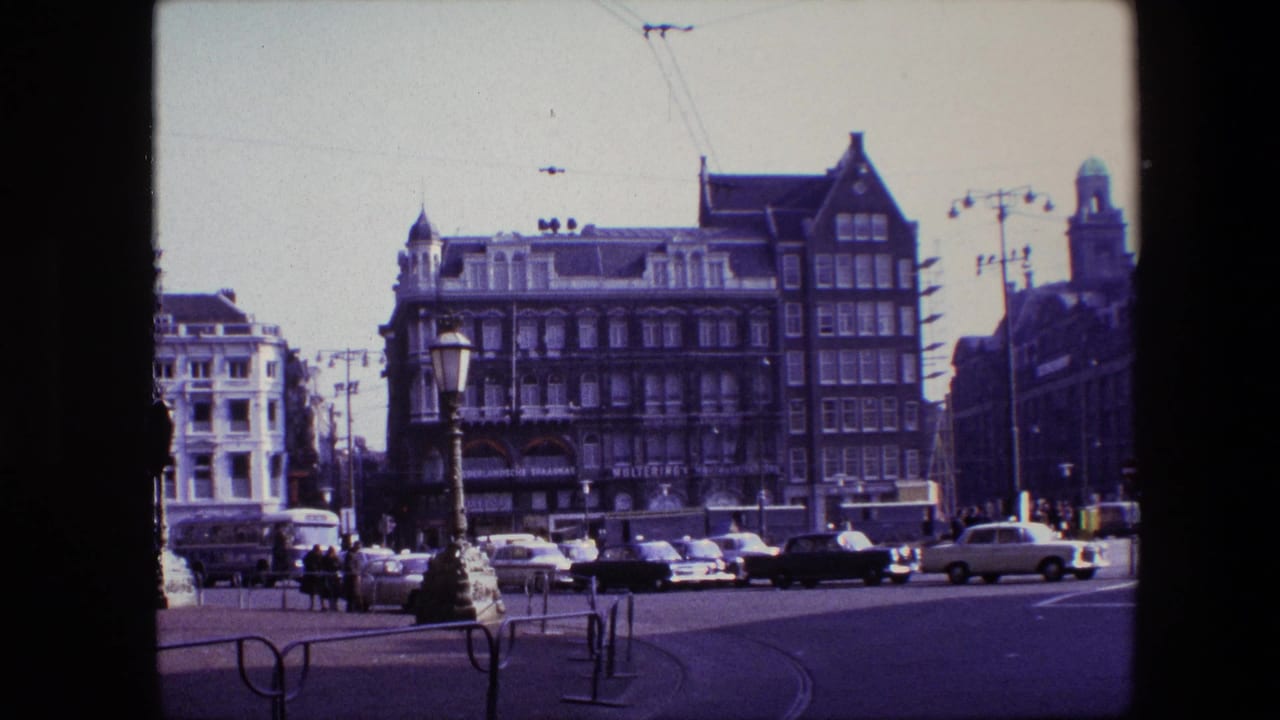 Traffic crosses an intersection with pedestrians near ornate buildings and tram wires overhead.