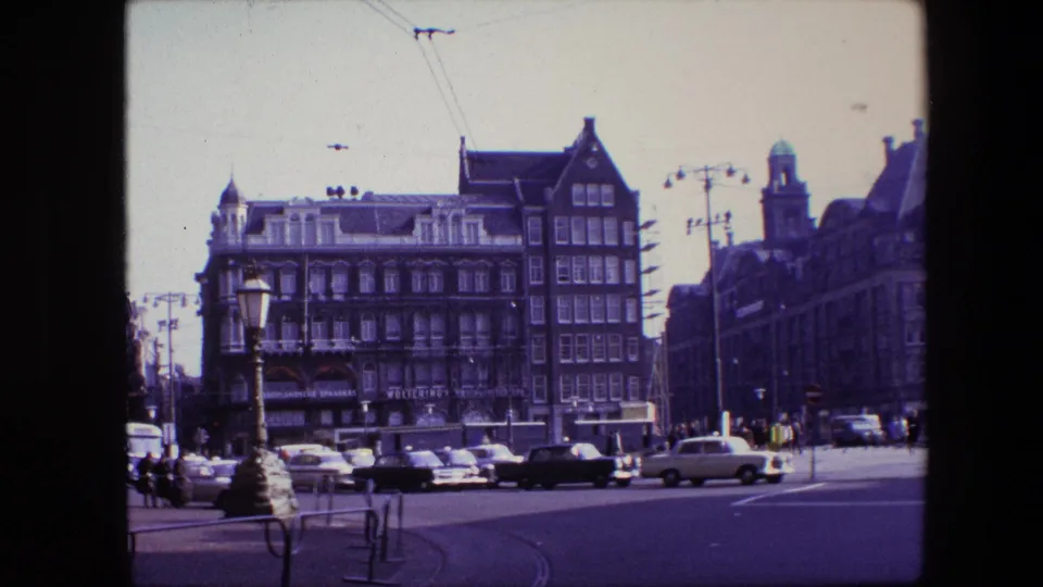 Stationary 8mm view of a 1967 Amsterdam intersection showing period cars, a light-colored vehicle, pedestrians, streetlamps, tram wiring, and tall gabled buildings.