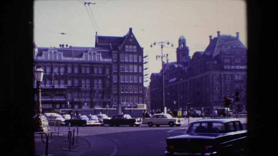 Amsterdam traffic scene from 1967 with a dark foreground car, other vehicles near the intersection, pedestrians, overhead tram wires, and commercial building signage in the background.