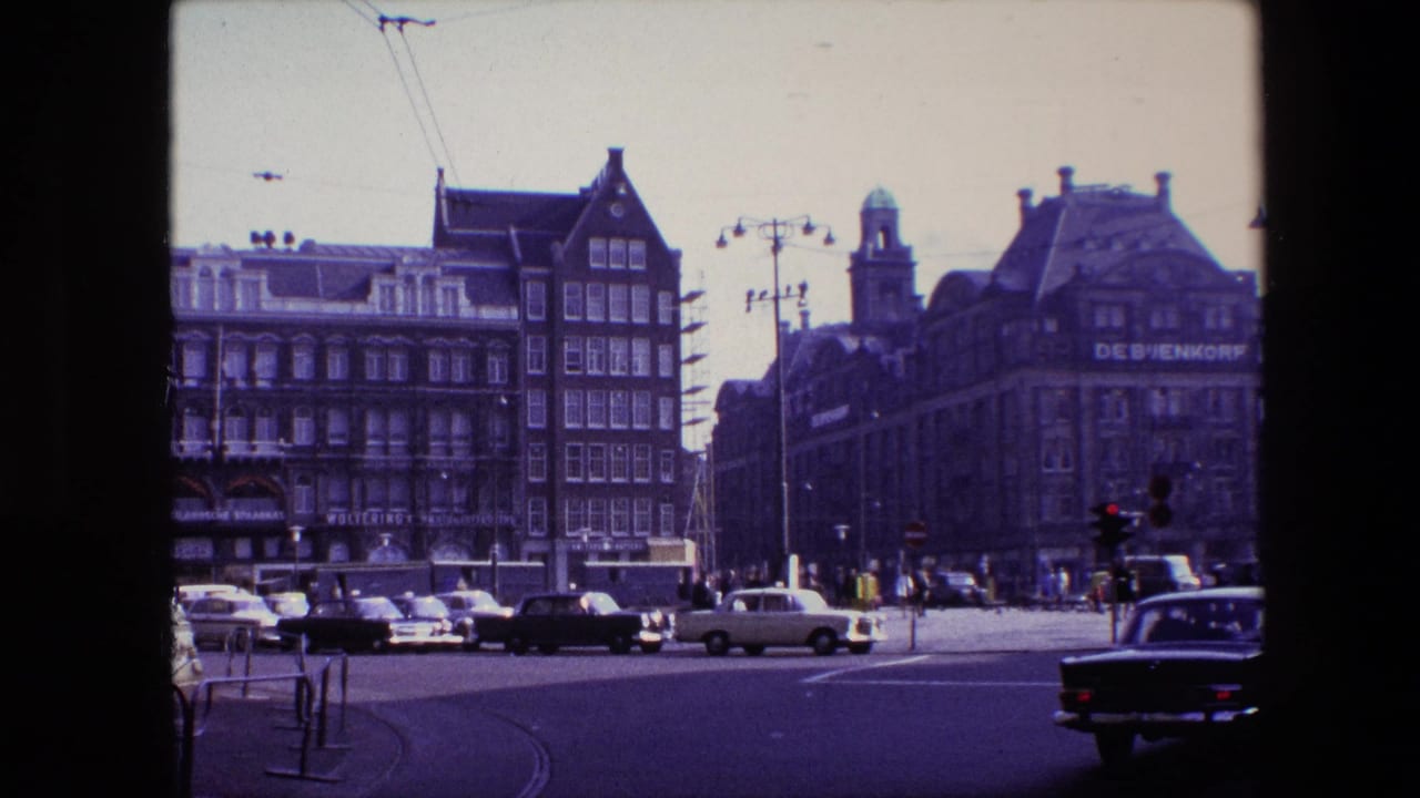 1967 Amsterdam street view showing classic cars moving through an intersection near the visible De Bijenkorf department store facade.