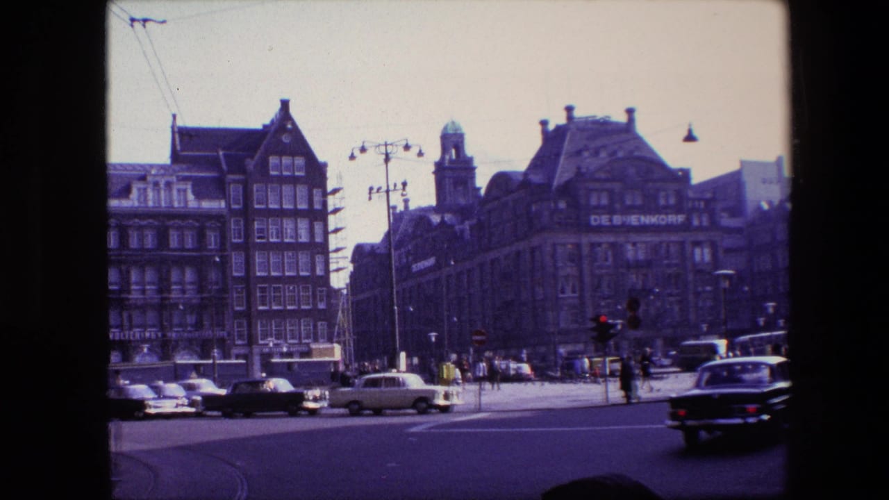 Amsterdam city intersection with mid-1960s cars passing in front of multi-story buildings and the De Bijenkorf sign visible in the background.