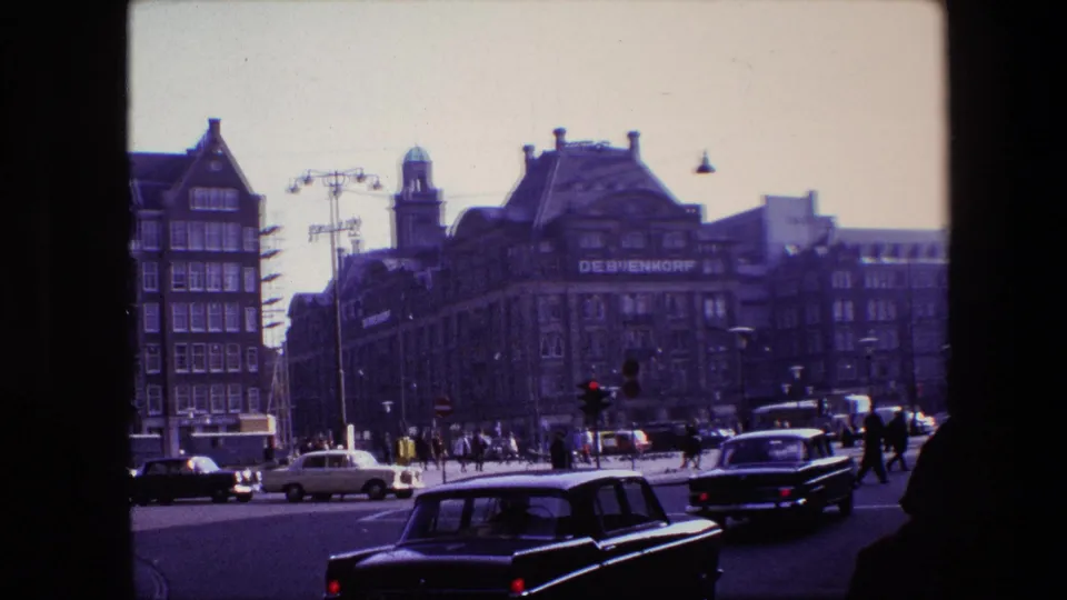 1967 Amsterdam home-movie frame with a dark car close to camera, additional traffic, pedestrians, and De Bijenkorf visible across the street.