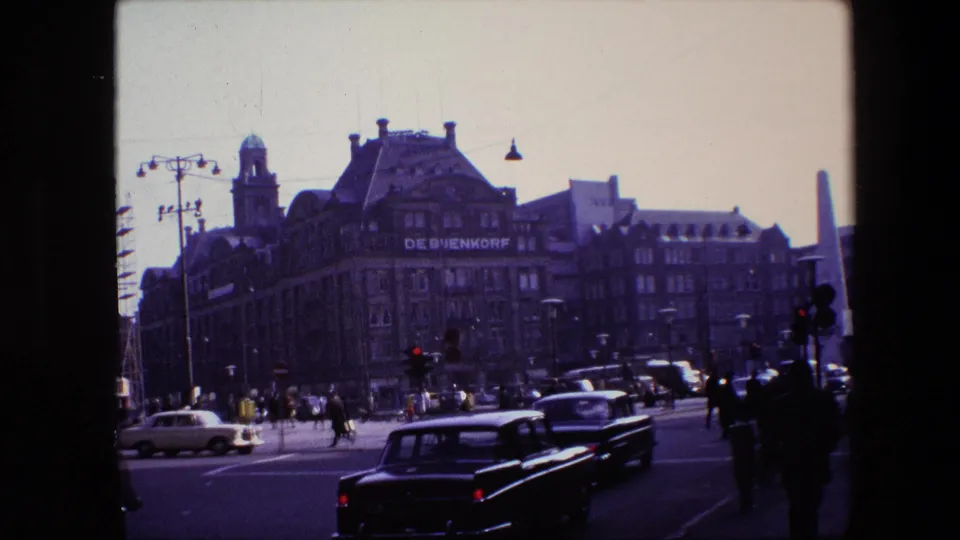 Amsterdam intersection in 1967 with classic cars, pedestrians, traffic lights, and the De Bijenkorf building seen through an 8mm home-movie frame.