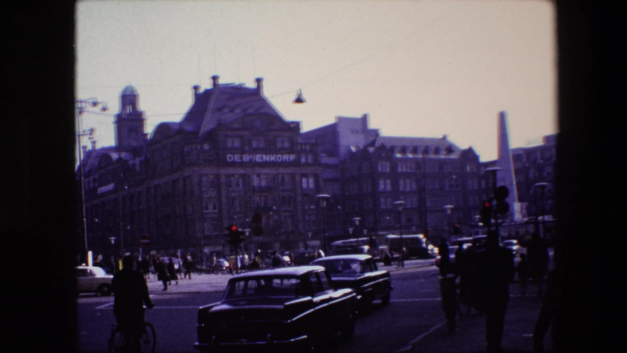 Amsterdam street view from 1967 metadata showing De Bijenkorf, classic cars, pedestrians, cyclists, traffic lights, and a tall monument in the distance.