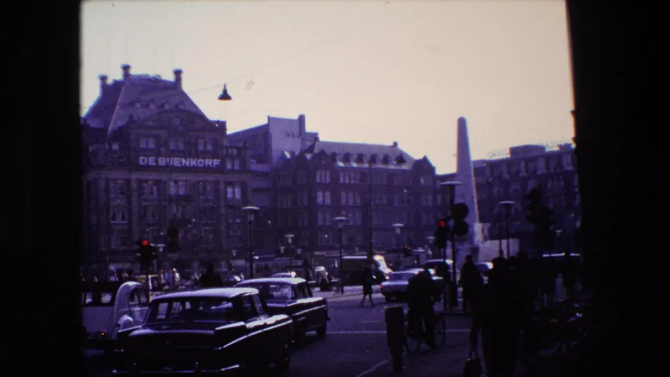 Amsterdam city-center street scene from the supplied 1967 clip metadata, with De Bijenkorf signage partly visible, classic cars, pedestrians, cyclists, and a tall monument beyond the roadway.