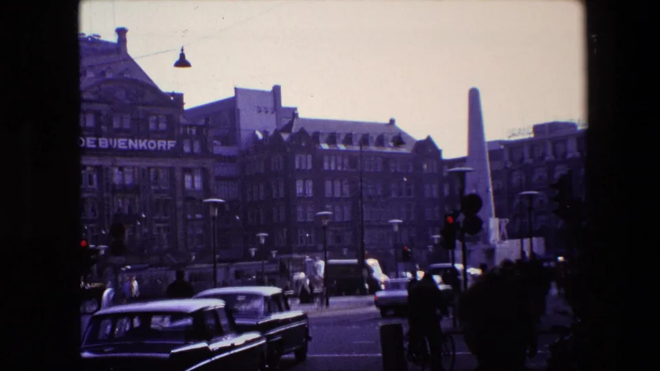 Late-1960s Amsterdam street view with a black car in the foreground, people moving near the crosswalk, De Bijenkorf signage on the building, and a tall monument in the background.