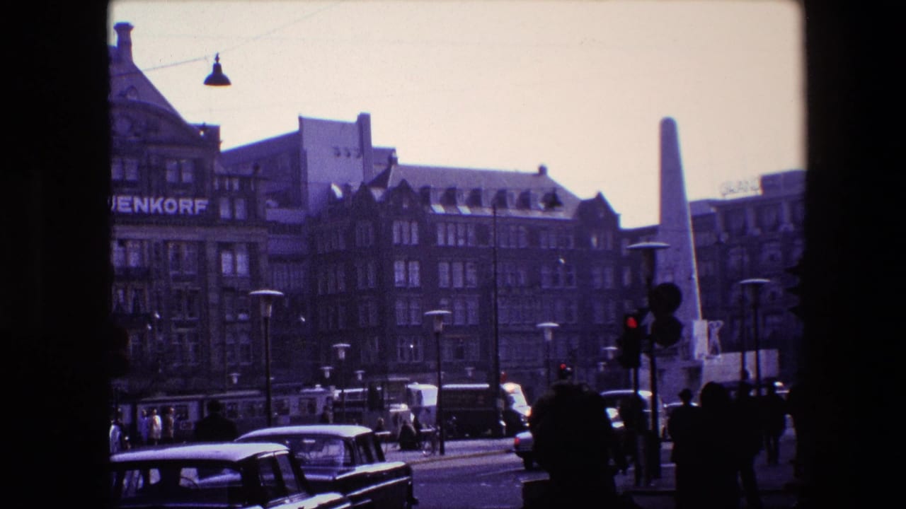 1967 Amsterdam street view showing cars and pedestrians near a tall obelisk-like monument, masonry buildings, traffic lights, and a partial commercial sign.