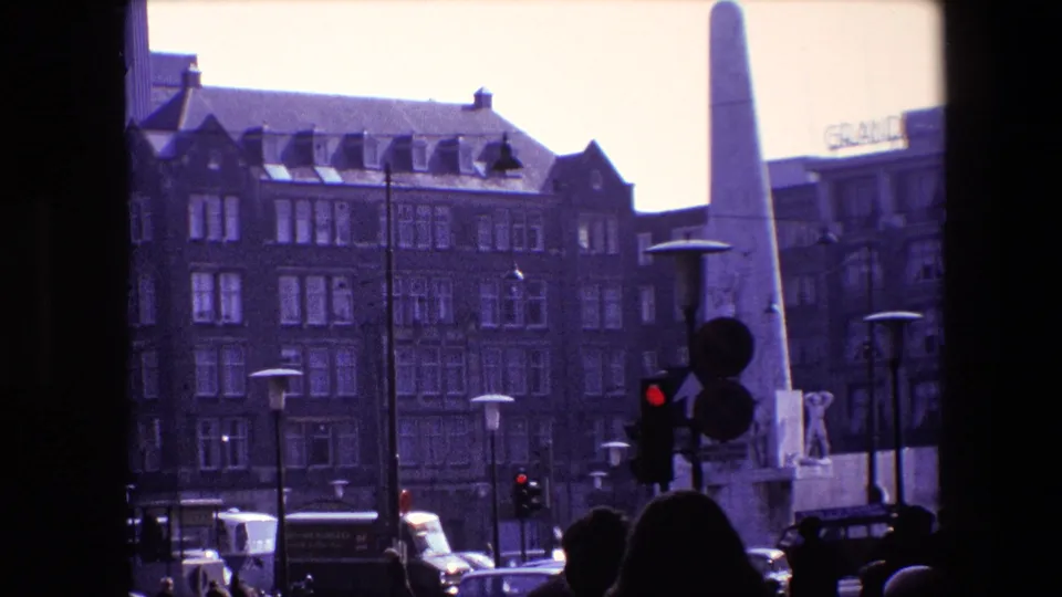 Closer 1967 Amsterdam view of the obelisk-like monument beside traffic lights, with pedestrians, cars, a bus or truck, and multi-story buildings behind it.