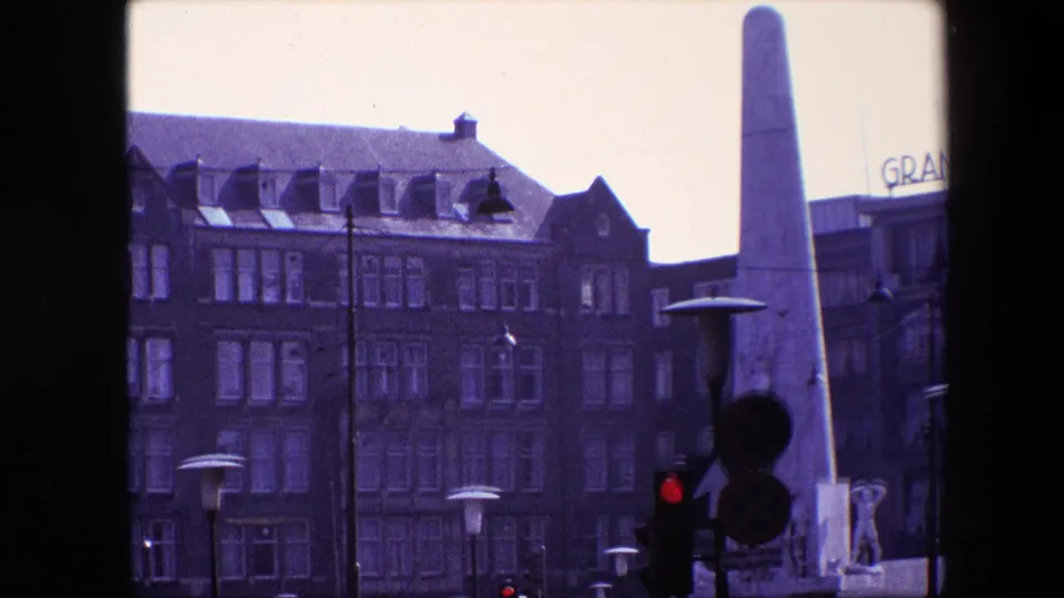 Amsterdam street frame showing the tall monument beside a traffic signal, with silhouettes of pedestrians, period cars, street lamps, and a partial sign on nearby buildings.