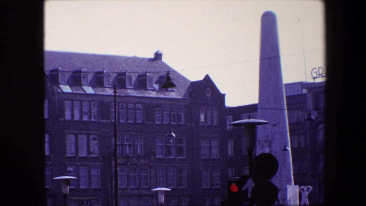 1967 Amsterdam street view with a pale obelisk, brick hotel-like facade, rooftop GRAND lettering, lamps, and traffic signal silhouettes.