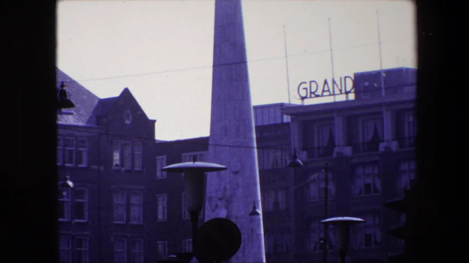 Amsterdam street frame showing the obelisk, dark brick architecture, rows of windows, lamps, traffic signal shapes, and visible GRAND sign lettering.