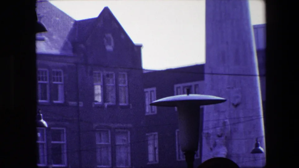 Final frame of the Amsterdam street scene, showing the tall obelisk, gabled brick facade, windows, hanging lamps, and a broad street lamp in the foreground.
