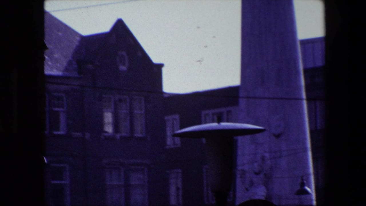 Amsterdam 1967 home-movie frame seen through a dark window or structural frame, with a building facade at left and the edge of a busy street visible beyond.
