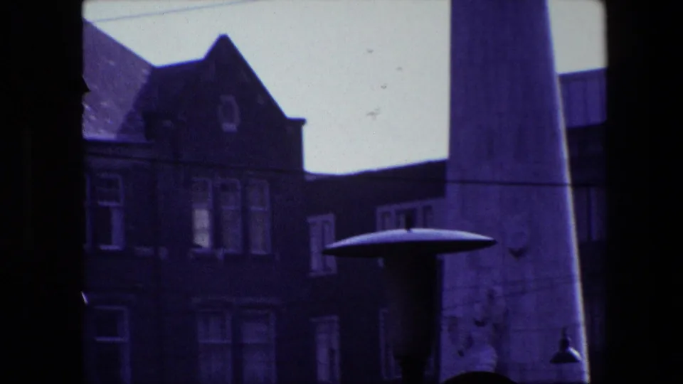 Amsterdam 1967 home-movie frame seen through a dark window or structural frame, with a building facade at left and the edge of a busy street visible beyond.